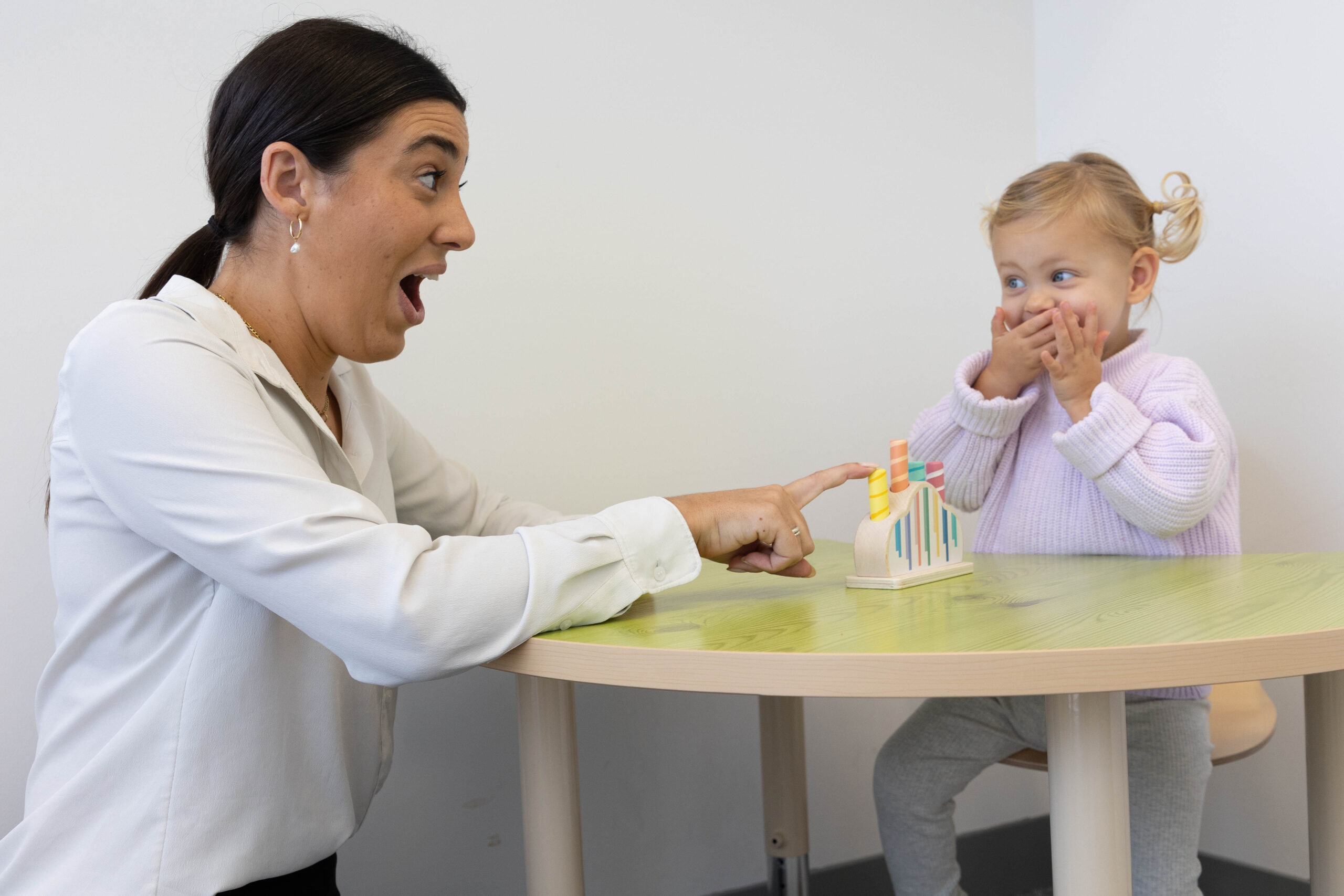 Woman and a child sit at a small table. Adult points to a toy while the child covers their mouth