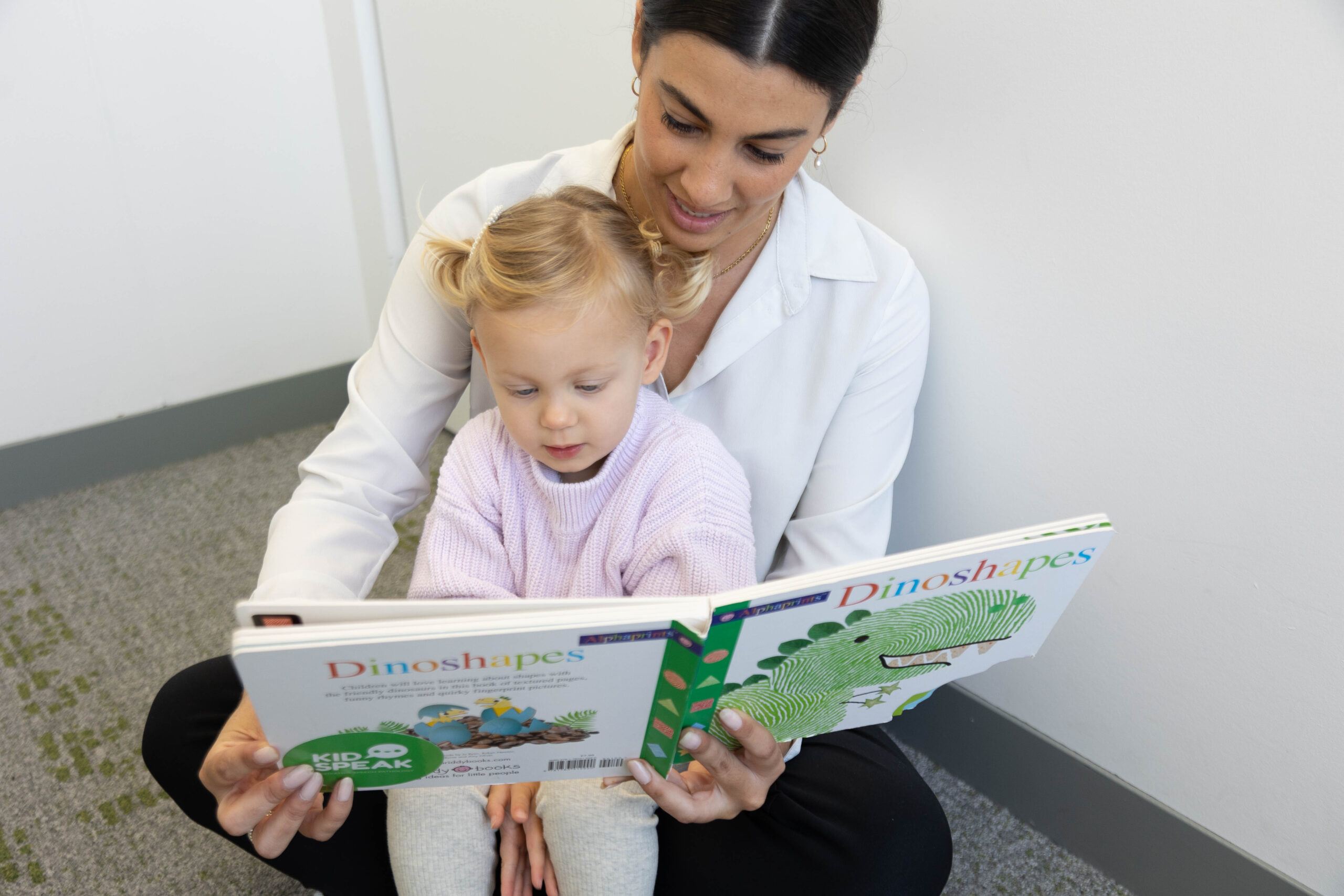 An adult and a child sit on the floor reading a book titled "Dinoshapes" together.