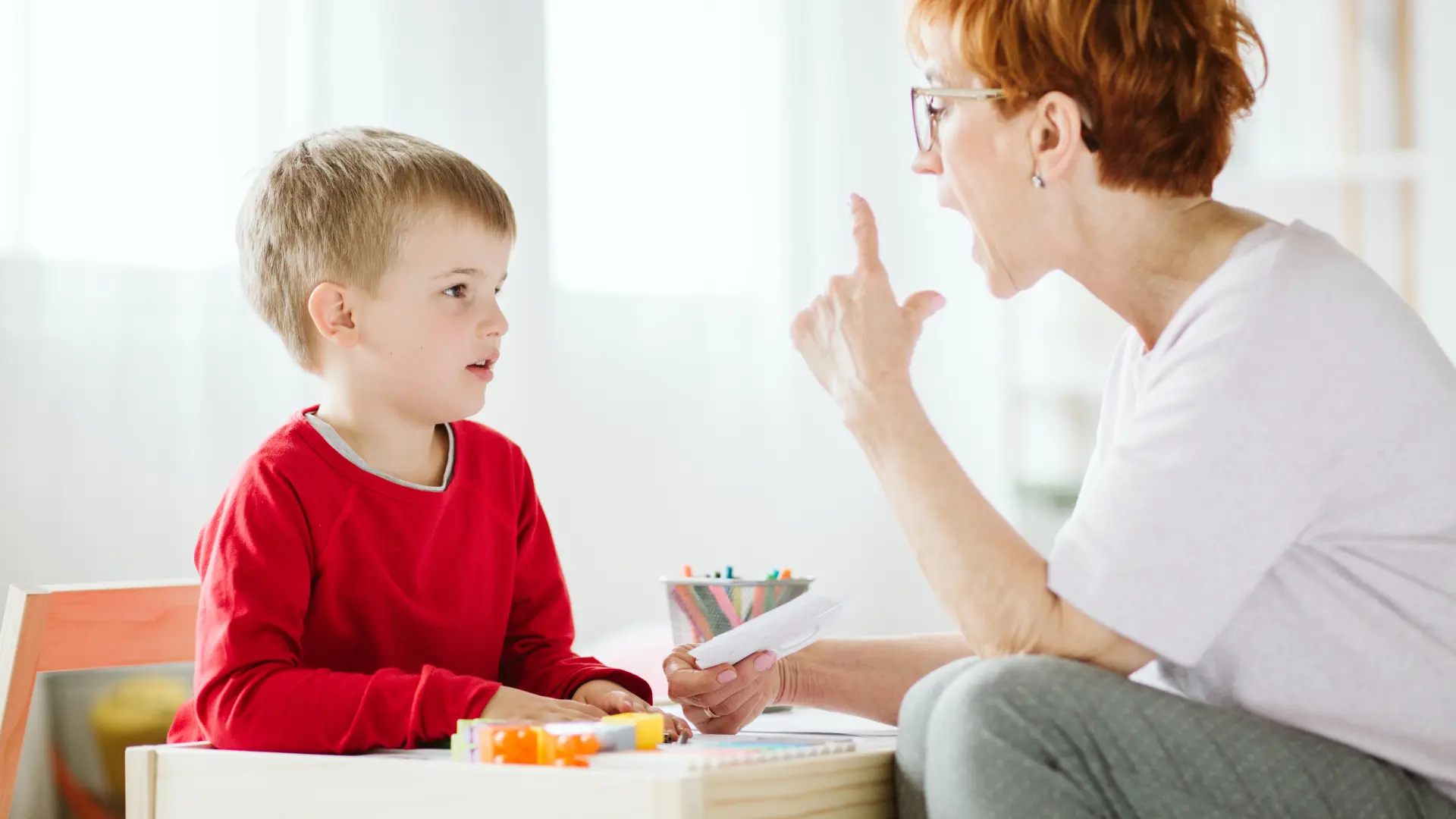 A young boy in a red shirt sits at a table during a speech therapy session